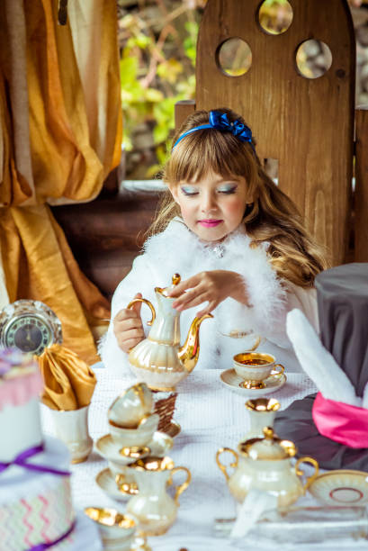 vista frontal de uma menina pouco bonita no cenário da alice no país das maravilhas, despejando um copo na mesa de chá - little girls alice in wonderland child fairy tale - fotografias e filmes do acervo