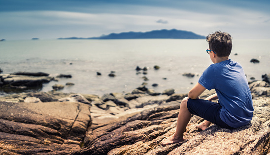 Cute boy sitting on rocks and watching at tranquil sea