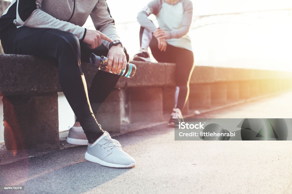 Couple of athletes resting after workout on the street Couple of athletes resting after workout session on the street. Tired man with bottle tracking time with his watch and woman relaxing on the bench Resting Stock Photo