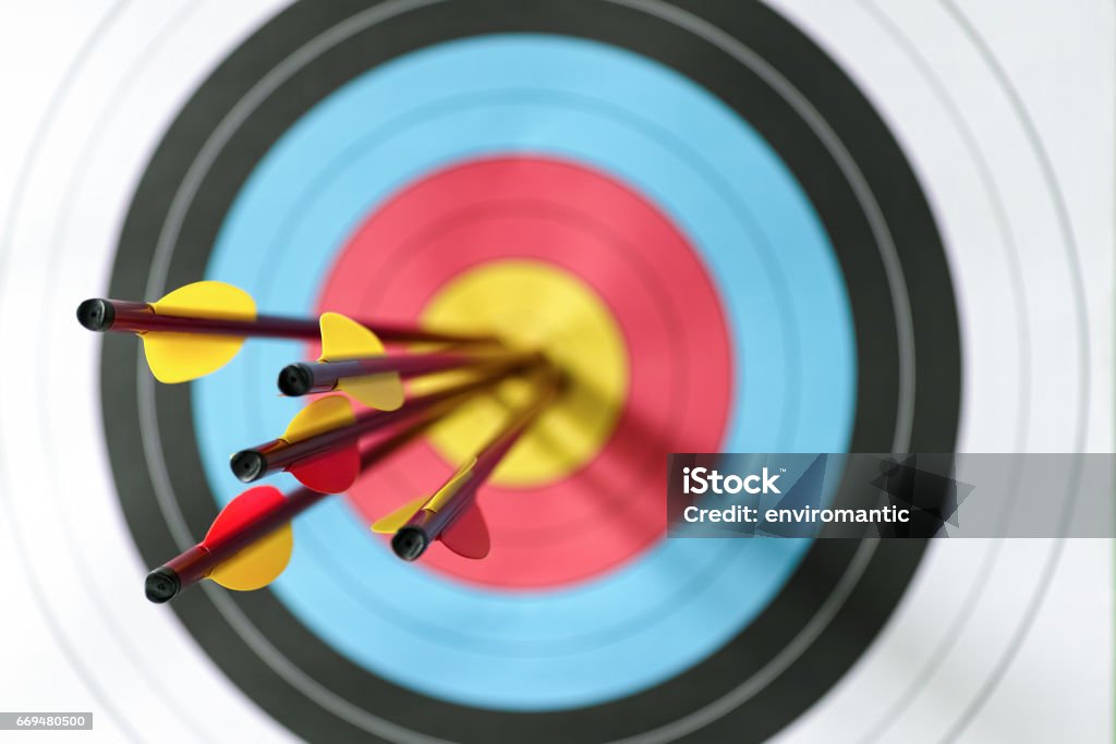 Five arrows in the bull's-eye of a sports target. Five arrows in the bull's-eye of an archery target. Selective focus with the focus being on the back end of the arrow, with an out of focus target in the background. Concept image being on target, strategy, aim, accomplishment, aiming for the bull's eye etc. Archery Stock Photo