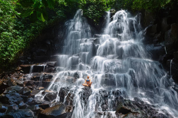 Woman sit on rock in yoga pose under cascade waterfall