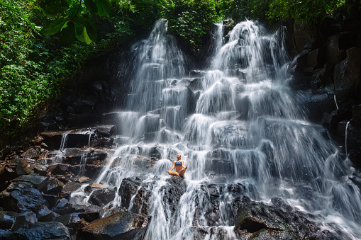 Travel in Bali jungle. Beautiful young woman sit in zen-like yoga pose under falling spring water, enjoy tropic cascade waterfall. Nature day trip, walking adventure, fun on family summer vacation