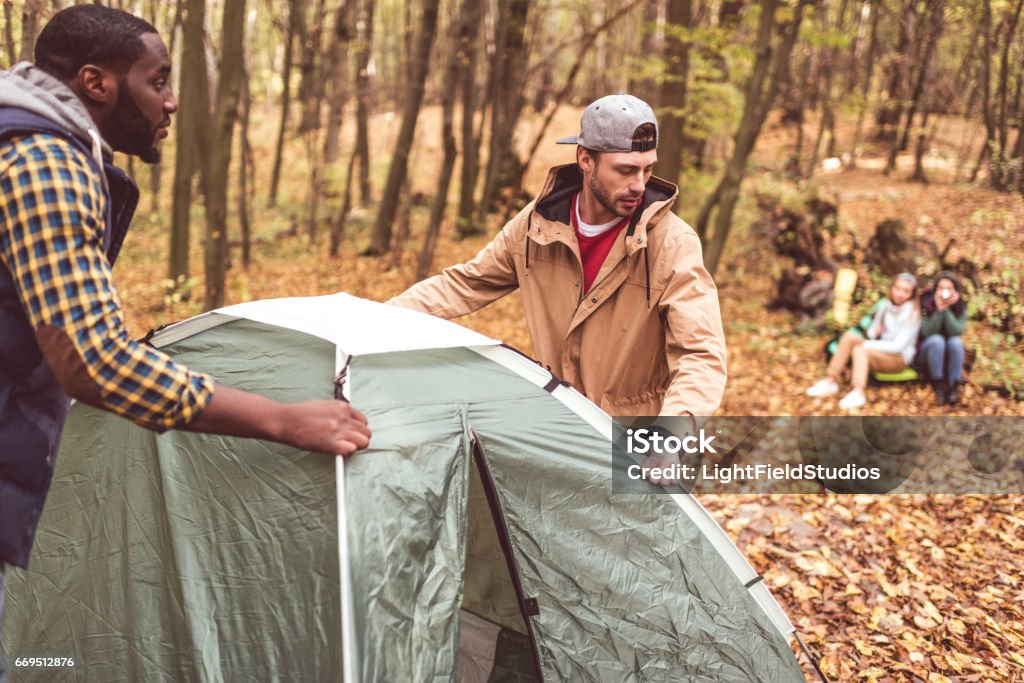 Men pitching tent in autumn forest Young men pitching tent in autumn forest with blurred girls sitting on background Tent Stock Photo