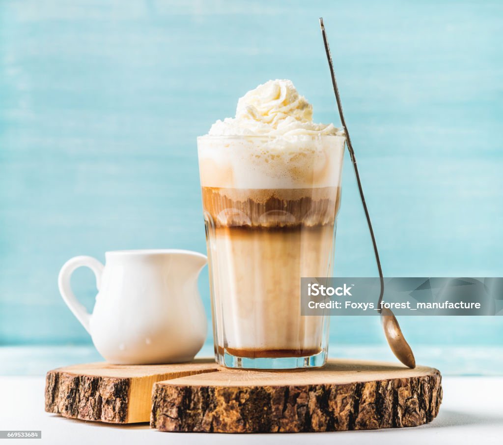 Latte macchiato with whipped cream, serving silver spoon and pitcher on wooden round board over blue painted wall background Latte macchiato with whipped cream, serving silver spoon and pitcher on wooden round board over blue painted wall background, selective focus, horizontal composition Latte Stock Photo