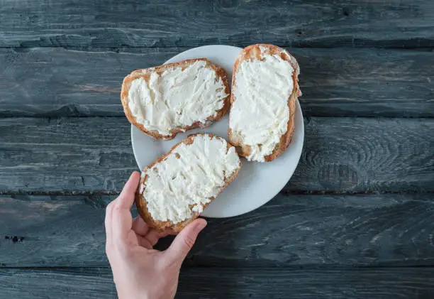 Hand holds homemade sandwich with cream-cheese over a black wooden table. Top view. Sandwiches on a white plate