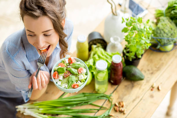 Woman eating healthy salad Young and happy woman eating healthy salad sitting on the table with green fresh ingredients indoors raw diet stock pictures, royalty-free photos & images