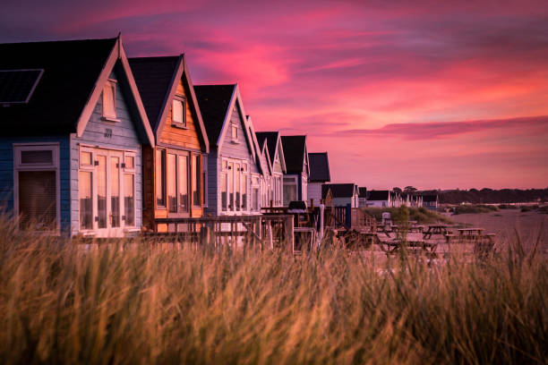 Hengistbury Head Beach Huts at Sunrise The sun rises in front of beach huts arranged along the sand spit at Hengistbury Head, Bournemouth. These solar powered huts are accessed via ferry from Mudeford or along a small road leading to Tuckton, at the edge of Bournemouth. They are among the most expensive beach huts in the United Kingdom and among the sand dunes which surround them lives the rare Sand Lizard. sandbar stock pictures, royalty-free photos & images