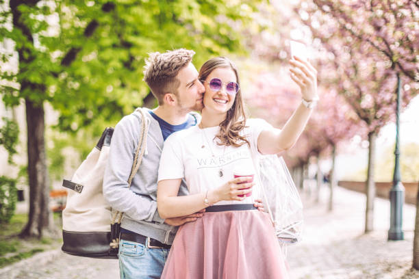Close-up of a blonde woman takes a photo of a cherry blossom on her phone. Woman in a peach sweater takes a photo of sakura.
