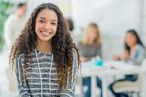 An girl of African descent is posing while inside a university campus. She has her arms crossed and is smiling at the camera. Her friends are studying in the background.