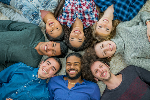 A multi-ethnic group of  university students are lying in a circle on the floor. They are looking up at the camera and smiling. They are wearing stylish, casual clothing.