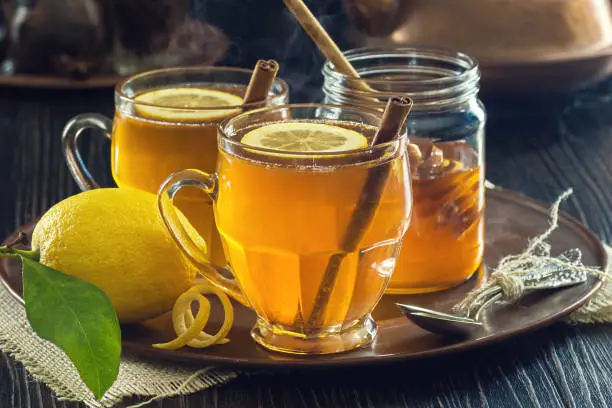 Two glasses of lemon spice tea or hot toddies with cinnamon sticks, lemon slices, and honey. The drinks are on a tray on a wooden table with a teapot and spices in the background.