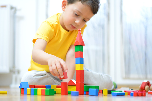 Happy mom and cheerful cute preschool kid girl playing on heating floor at home, stacking colorful cubes, constructing toy towers from building blocks, talking, laughing, smiling, enjoying leisure