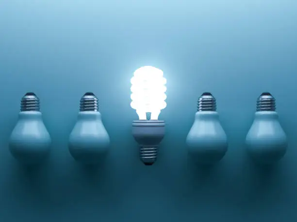 Photo of Energy saving light bulb up , one glowing fluorescent lightbulb standing out from unlit down incandescent bulbs on blue background , individuality and different creative idea concepts . 3D rendering