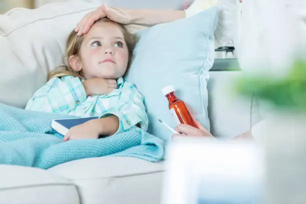 Young Caucasian girl is ill with a cold or a flu virus. Her mother or grandmother is checking her forehead to see if she has a fever. The caretaker is holding a bottle of medicine.