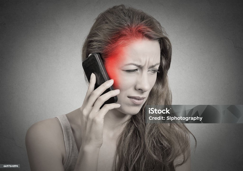 girl on the phone with headache. Upset unhappy female talking on phone girl on the phone with headache. Upset unhappy female talking on phone isolated grey wall background. Negative human emotion face expression feeling life reaction. Cellular mobile radiation concept Adult Stock Photo