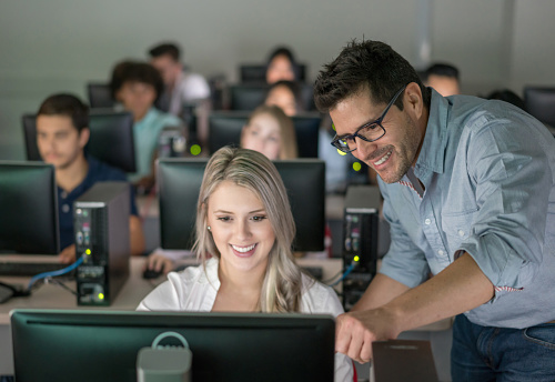 Teacher helping a female student at an IT class pointing the screen of her computer and looking happy - education concepts