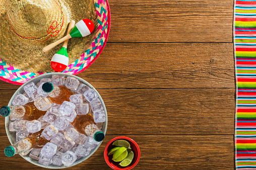 A Mexican theme celebration viewed from overhead. Included is a bucket of beer bottles on ice, a bowl of limes, a sombrero, maracas and a multi-colored striped blanket sitting on some brown wooden planks. This could be for Cinco de Mayo or any other Mexican celebration of holiday.
