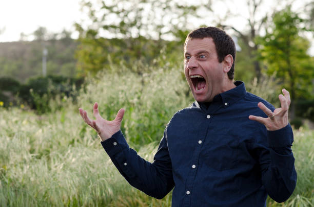 Man screaming in anger with hands out and mouth wide open outside in a grassy field wearing a long sleeve button up shirt.