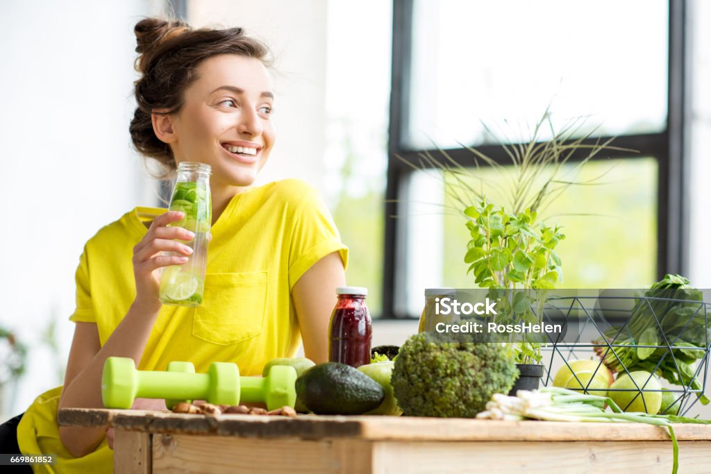 Woman with healthy food indoors Portrait of a young sports woman in yellow t-shirt sitting indoors with healthy food and dumbbells on the table Drinking Stock Photo