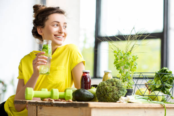 Woman with healthy food indoors Portrait of a young sports woman in yellow t-shirt sitting indoors with healthy food and dumbbells on the table detox stock pictures, royalty-free photos & images