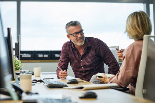 Businessman communicating with colleague at desk Smiling businessman communicating with colleague at desk. Male and female professionals are sitting in office. They are discussing strategies at textile industry. man beard plaid shirt stock pictures, royalty-free photos & images