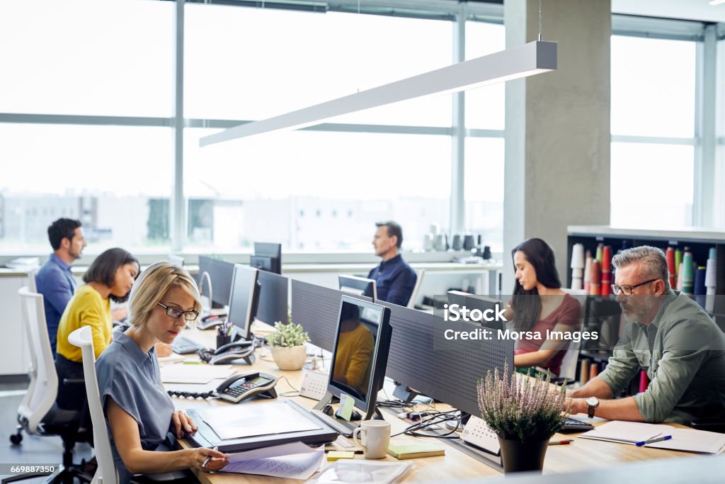 Business people working at desk by windows Business people working at desk. Male and female professionals are sitting by windows. Colleagues are sitting in textile industry. Office Stock Photo
