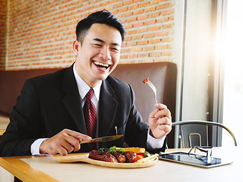 Happy young businessman eating rib steak on wooden tray at restaurant. Copy space.