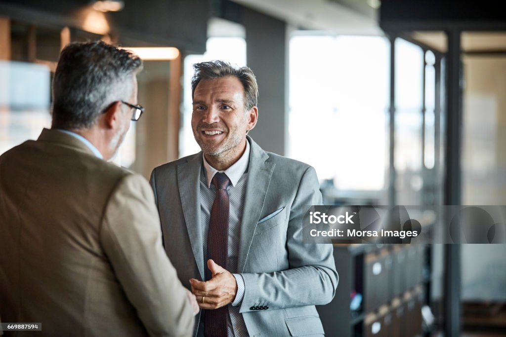 Businessmen communicating in textile factory Senior businessman smiling while looking at colleague in textile factory. Professional is wearing suit while looking at coworker. They are communicating in office. Discussion Stock Photo