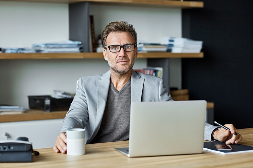 Portrait of confident businessman with laptop at desk. Senior executive is sitting in office. He is wearing smart casual at textile factory.