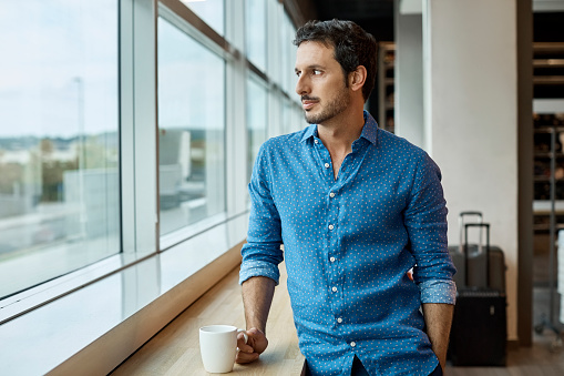 Thoughtful businessman holding coffee cup at window sill. Male professional is looking through window. He is standing in textile factory.