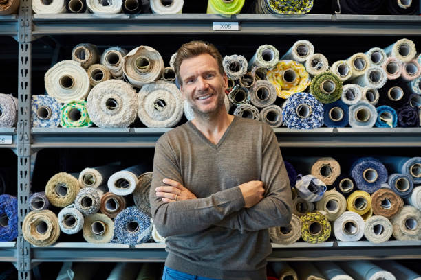 Executive with arms crossed against rolled fabric Portrait of confident businessman with arms crossed against rolled fabric. Executive is smiling while standing against shelves. He is wearing smart casuals in textile factory. textile industry stock pictures, royalty-free photos & images