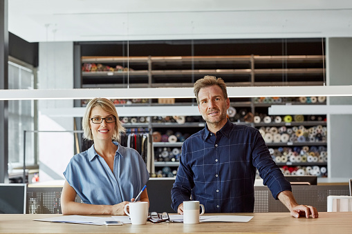 Portrait of confident colleagues smiling at desk. Business professionals are standing in textile factory. They are wearing smart casuals.