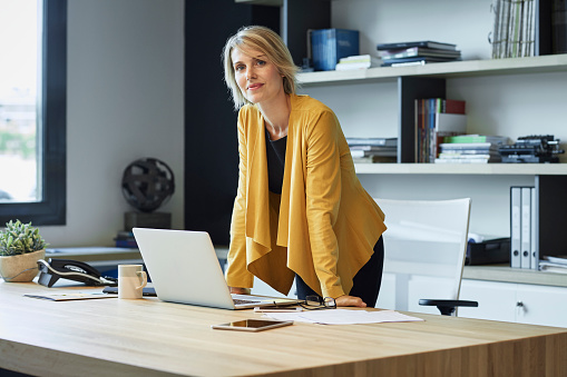 Portrait of confident businesswoman leaning on desk. Female professional is with laptop. She is standing in textile industry.