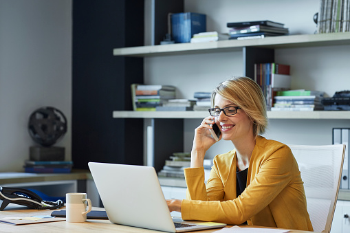 Businesswoman using laptop and smart phone at desk. Confident female professional is working in textile factory. Executive is smiling in office.