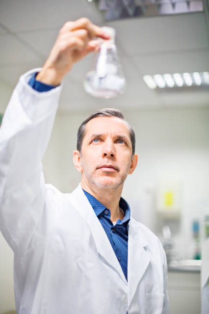 Male scientist examining chemical solution in flask at laboratory. Professional is performing experiment at pharmacy. He is wearing lab coat.
