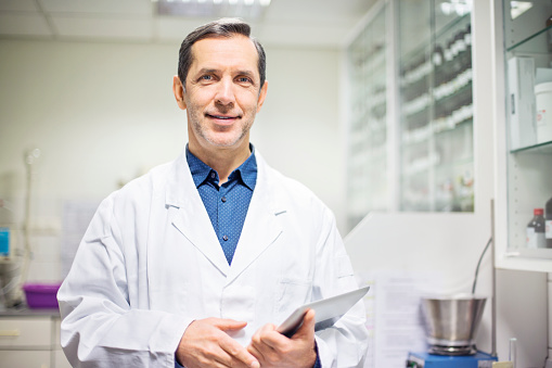 Portrait of happy male scientist holding digital tablet in laboratory. Smiling doctor is standing at pharmacy. He is wearing lab coat.