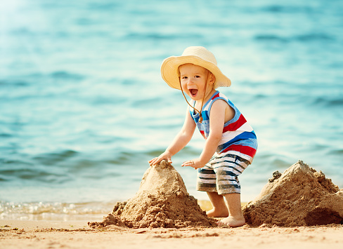 one year old boy walking at the beach in straw hat. Child on family vacations at sea building a castle
