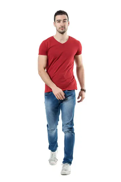 Relaxed casual man in jeans and red t-shirt walking and looking at camera. Full body length portrait isolated over white studio background.