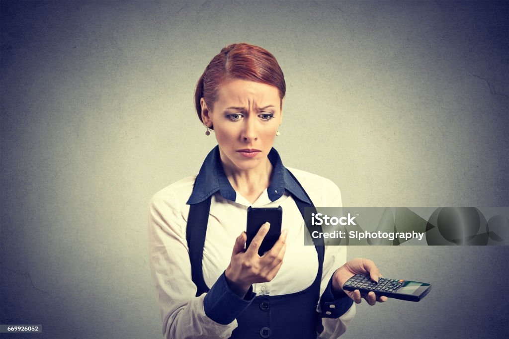 Sad unhappy upset business woman looking at smart phone holding calculator unexpected financial bills charges Sad unhappy upset business woman looking at smart phone holding calculator unexpected financial bills charges isolated on gray wall background. Human face expression, emotion, body language, reaction Adult Stock Photo