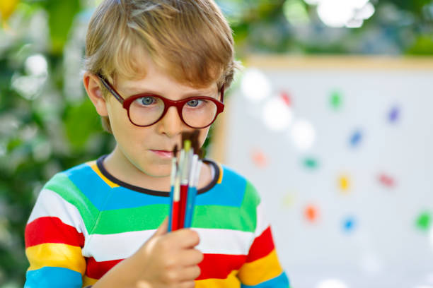 Confused little kid boy with glasses holding watercolors and brushes. Happy child and student is back to school. Education, school, learning concept. School, preschool nursery equipment
