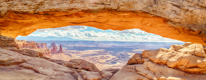 Panoramic view of famous Mesa Arch, iconic symbol of the American West, illuminated golden in beautiful morning light on a sunny day with blue sky and clouds, Canyonlands National Park, Utah, USA