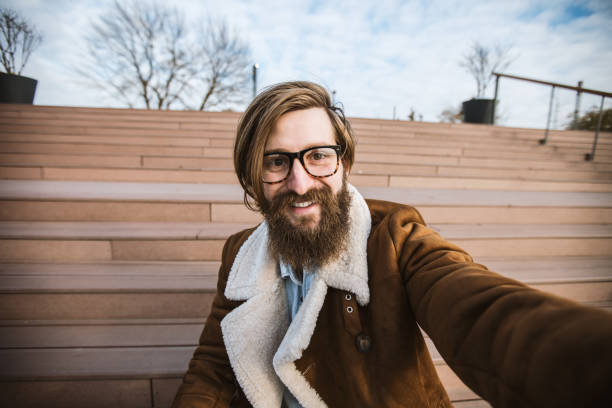 long-haired berded guy sitting on the staiway taking photo of himself. selfie - stubble men tattoo sensuality imagens e fotografias de stock