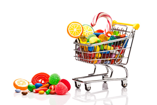 Front view of a miniature shopping cart filled with colorful candies, jellybeans and lollipops isolated on reflective white background. Some candies are out of the shopping cart. Copy space available at the left of the fram. DSRL studio photo taken with Canon EOS 5D Mk II and Canon EF 100mm f/2.8L Macro IS USM