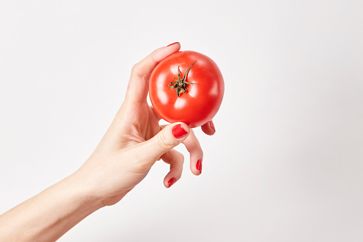 Fresh vegetable tomato in woman hand, fingers with red nails manicure, isolated on white background, healthy lifestyle concept