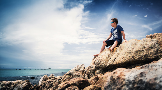 Cute boy sitting on rocks and watching at tranquil sea