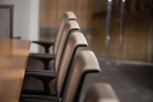 Modern conference room with long wooden table and lime grey office chairs lined up around it. Walls of glass and light shine through.