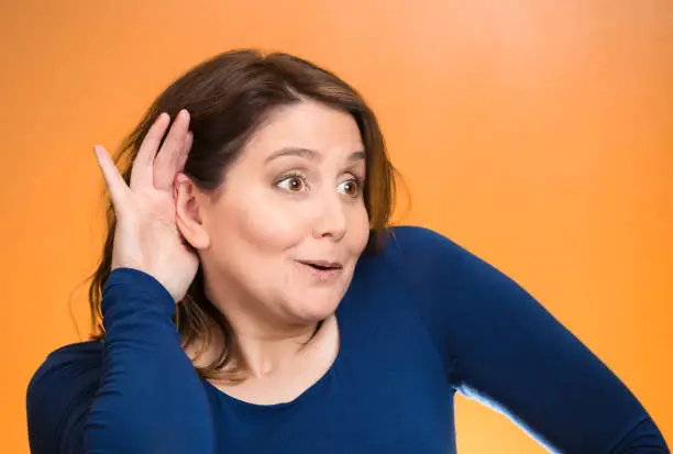 Closeup portrait curious, nosy woman listening to someone's conversation, hand to ear gesture,  looking surprised shocked by what she discovered isolated orange background. Human emotion expression