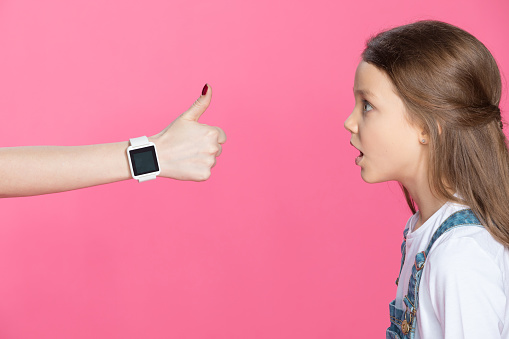 Side view of shocked little girl looking at woman with smartwatch showing thumb up