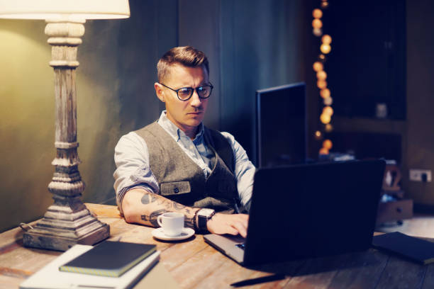 Elegant tattooed man working at home on laptop while sitting at the wooden table.Using modern computer for research new startup ideas.Concept of business people work.Blurred background hombre tatuado elegante trabajo en casa en la computadora portátil mientras está sentado en la mesa de madera. utilizando equipo moderno para las nuevas ideas de inicio investigación. concepto de trabajo de la gente de negocios. fondo borroso.