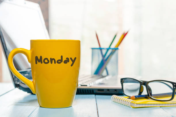 Monday written on yellow coffee or tea cup at wooden boards table, workplace, office sunlight morning background.